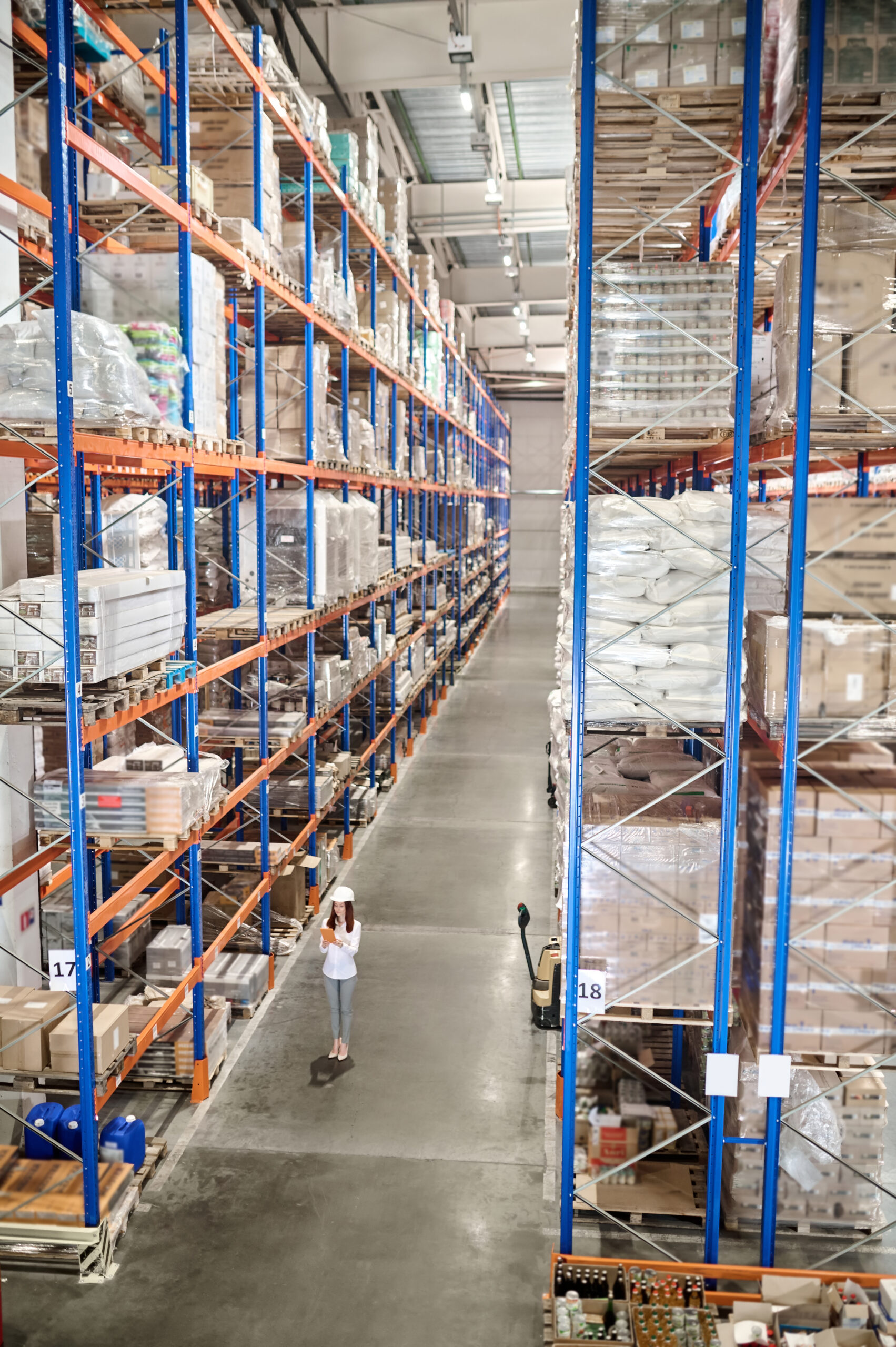 Large warehouse. Woman in white protective helmet with tablet standing in large industrial warehouse in aisle between high racks for goods